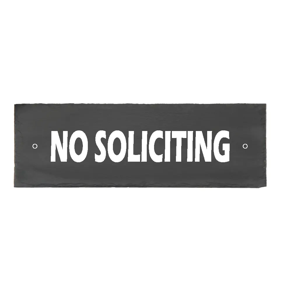 A rectangular 4x12 black slate No Soliciting sign with hand-chiseled edges and crisp white painted lettering, featuring pre-drilled mounting holes on a rustic wood background.