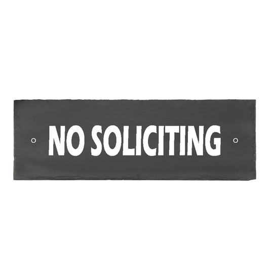 A rectangular 4x12 black slate No Soliciting sign with hand-chiseled edges and crisp white painted lettering, featuring pre-drilled mounting holes on a rustic wood background.